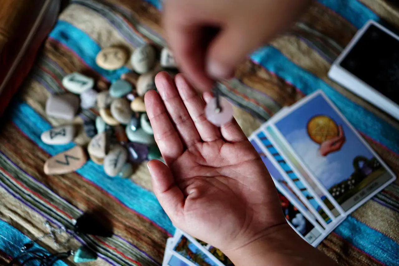 Hand holds a pendulum over runes and tarot cards, exploring divination.