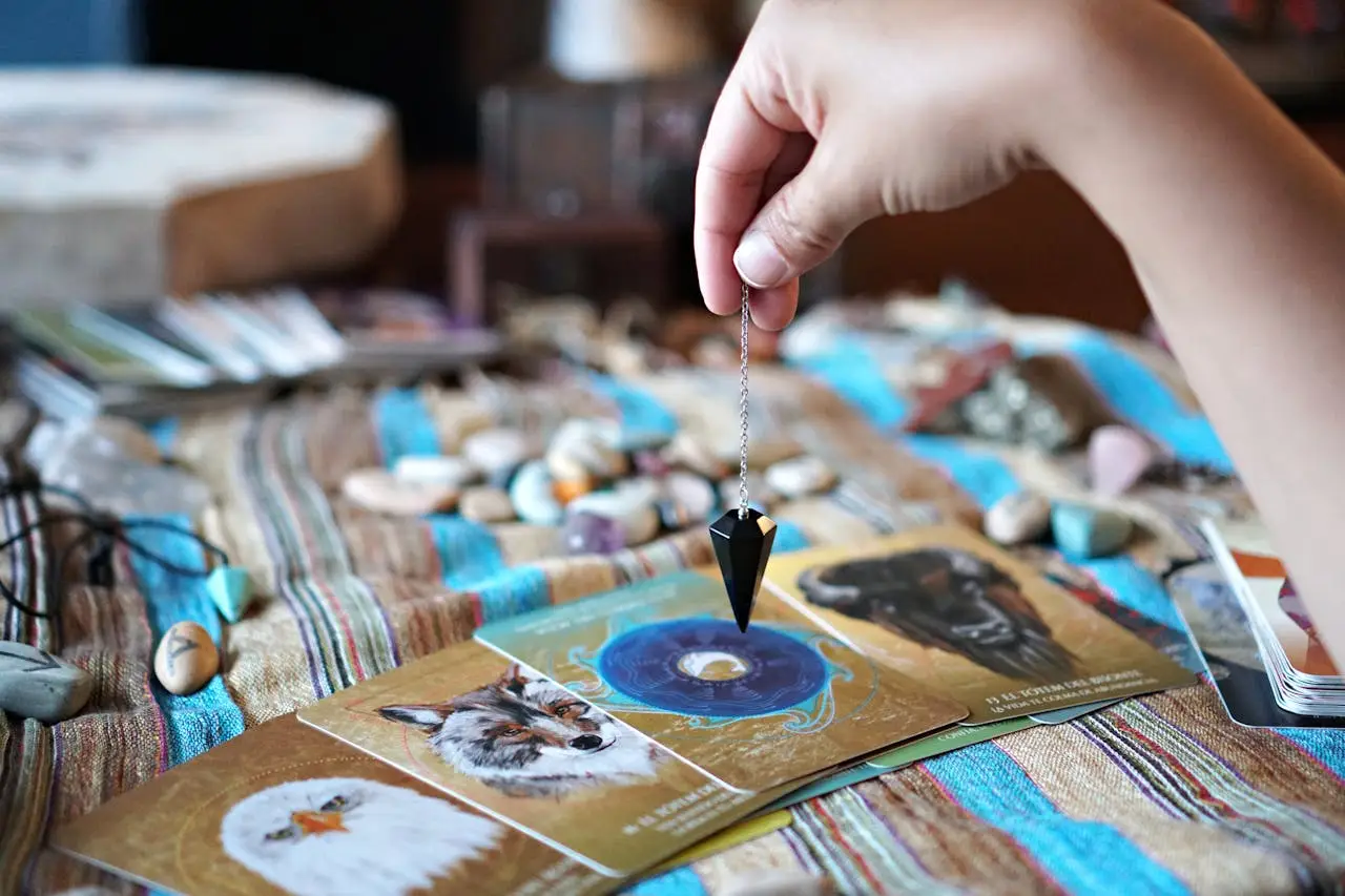 A hand holding a pendulum over tarot cards on a table with crystals and stones.